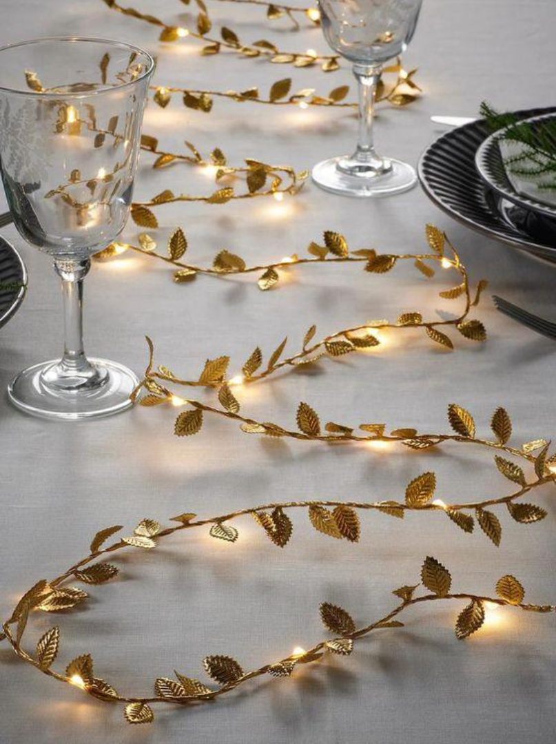 Goldene Lichterranke mit Blättern als Tischdeko auf weißer Tischdecke zwischen Weingläsern und Tellern