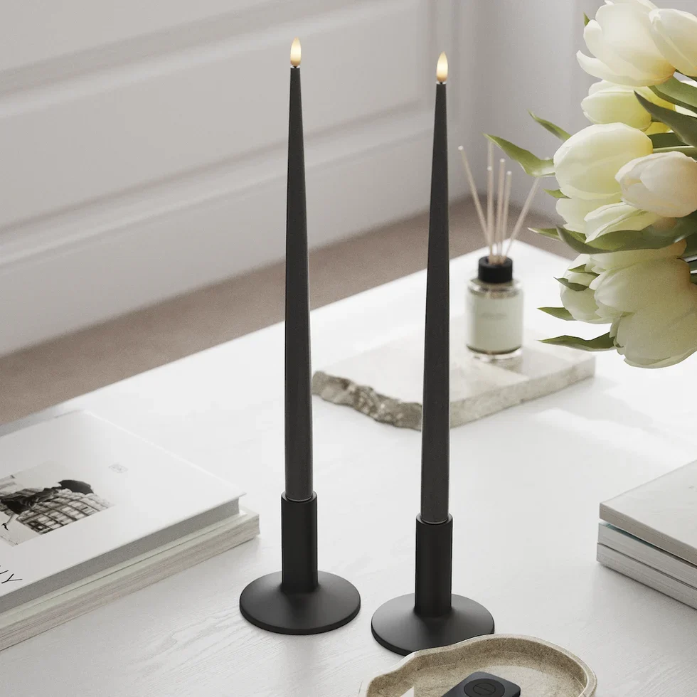 Zwei graue LED-Stabkerzen in schwarzen Kerzenhaltern auf weißem Couchtisch mit Büchern, Tulpen und Duftstäbchen