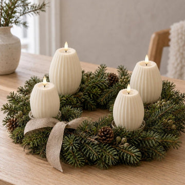 Vier cremefarbene gerillte LED-Kerzen in bauchiger Form in zwei Größen in einem Adventskranz aus Tannengrün mit Zapfen und Leinenschleife auf einem Holztisch