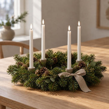 Vier weiße LED-Stabkerzen im Wetlook in einem Adventskranz aus Tannengrün mit Zapfen und Leinenschleife auf einem Holztisch