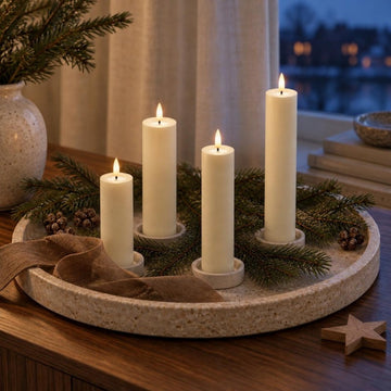 Vier schlanke cremefarbene LED-Stumpenkerzen in vier Höhen auf einem runden Terrazzo-Tablett mit Tannengrün und Band auf einem Sideboard am Winterabend