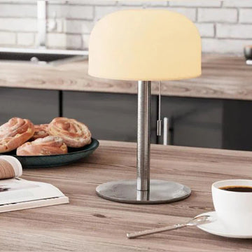 Eingeschaltete graue Tischlampe mit warmem Licht auf Küchentheke mit Kaffee, Zimtschnecken und Magazin