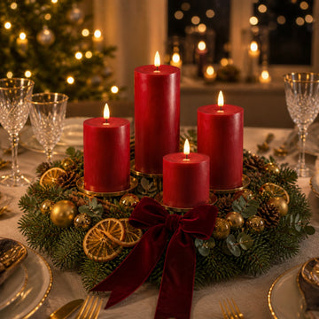 Vier bordeauxrote LED-Stumpenkerzen in einem Adventskranz aus Tannengrün mit Goldkugeln, Orangenscheiben und roter Samtschleife auf der Festtafel